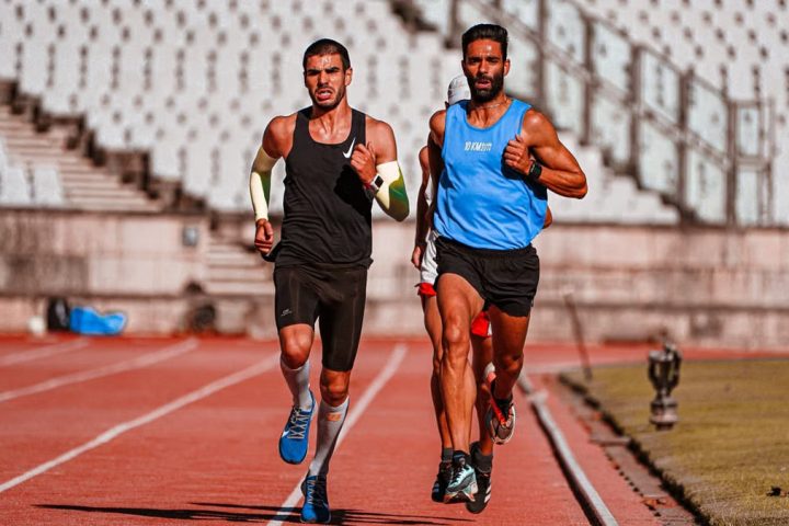 Two male runners competing on a stadium track during a sunny day.
